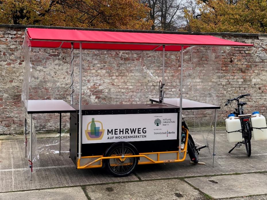 Ein zum Marktstand erweitertes Lastenrad mit der Aufschrift: Mehrweg auf Wochenmärkten.