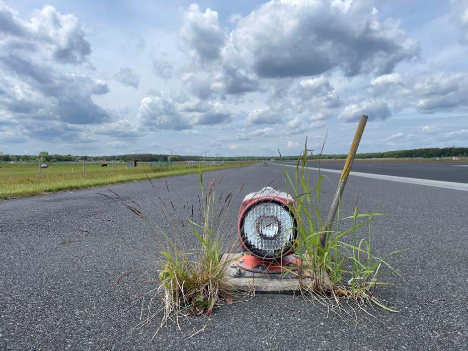 Start- und Landebahn des Flughafens Tegel aus der Froschperspektive, ganz vorn eine schon mit etwas Gras bewachsene runde Leuchte, ganz hinten erstreckt sich die Startbahn bis zum Horizont.