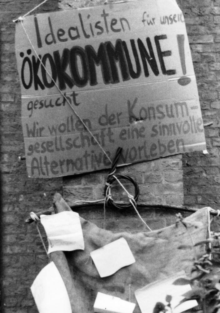 Ein handgemaltes Pappschild mit der Aufschrift: Idealisten für unsere Öko-Kommune gesucht! Wir wollen der Konsumgesellschaft eine sinnvolle Alternative vorleben.