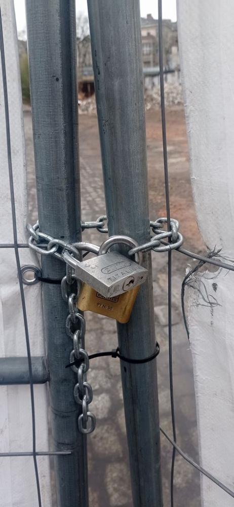 Nahaufnahme: Zwei Bauzaun-Elemente, offenbar das Tor zum Baugelände, sind mit einer Kette und einem Vorhängeschloss verschlossen.