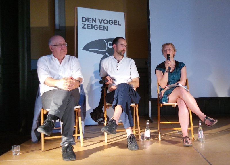 Bei einer Podiumsdiskussion sitzen ein älterer und ein junger Mann sowie eine junge Frau vorn, die Frau spricht ins Mikrofon. Hinten ist eine  Tafel mit dem Titel der Rabe-Ralf-Ausstellung zu sehen: Den Vogel zeigen.
