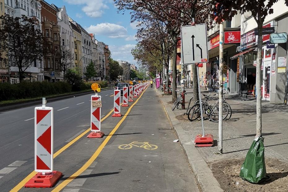 Am Rand einer Berliner Hauptstraße ist eine Fahrspur mit mobilen Baken und zwei gelben Streifen abgetrennt und mit einem gelben Fahrrad als geschützter Radstreifen markiert.