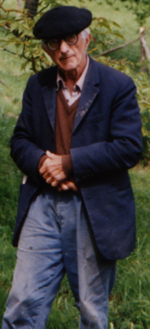 Zu sehn ist Bernard Charbonneau. Er steht auf einer Grünfläche und hat einen flachen Hut, eine blaue Anzugsjacke, einen braunen Pulli und eine hellblaue Jeans an.