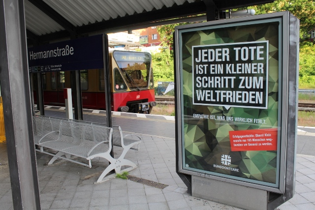 Zu sehen ist der S-Bahnhof Hermannstraße mit einer Sitzbank, Reklametafel und einer S-Bahn im Hintergrund.