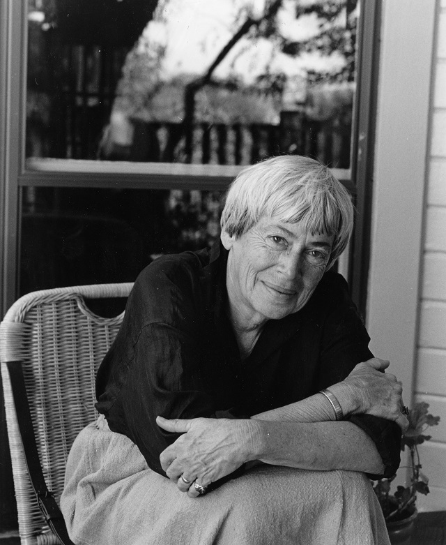 Zu sehen ist ein schwarz-weiß Foto von Ursula K. Le Guin. Sie sitzt auf einem Stuhl und hat einen dunklen Pulli und eine helle Hose an. Sie hat kurze weiße Haare.