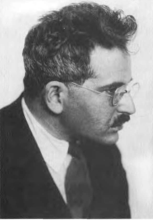 Mna kann ein schwarz-weiß Foto von Walter Benjamin sehen. Man hat eine seitliche Sicht auf ihn. Er hat buschiges dunkles Haar, eine Brille, einen Schnurrbart und einen Anzuf mit Krawatte.